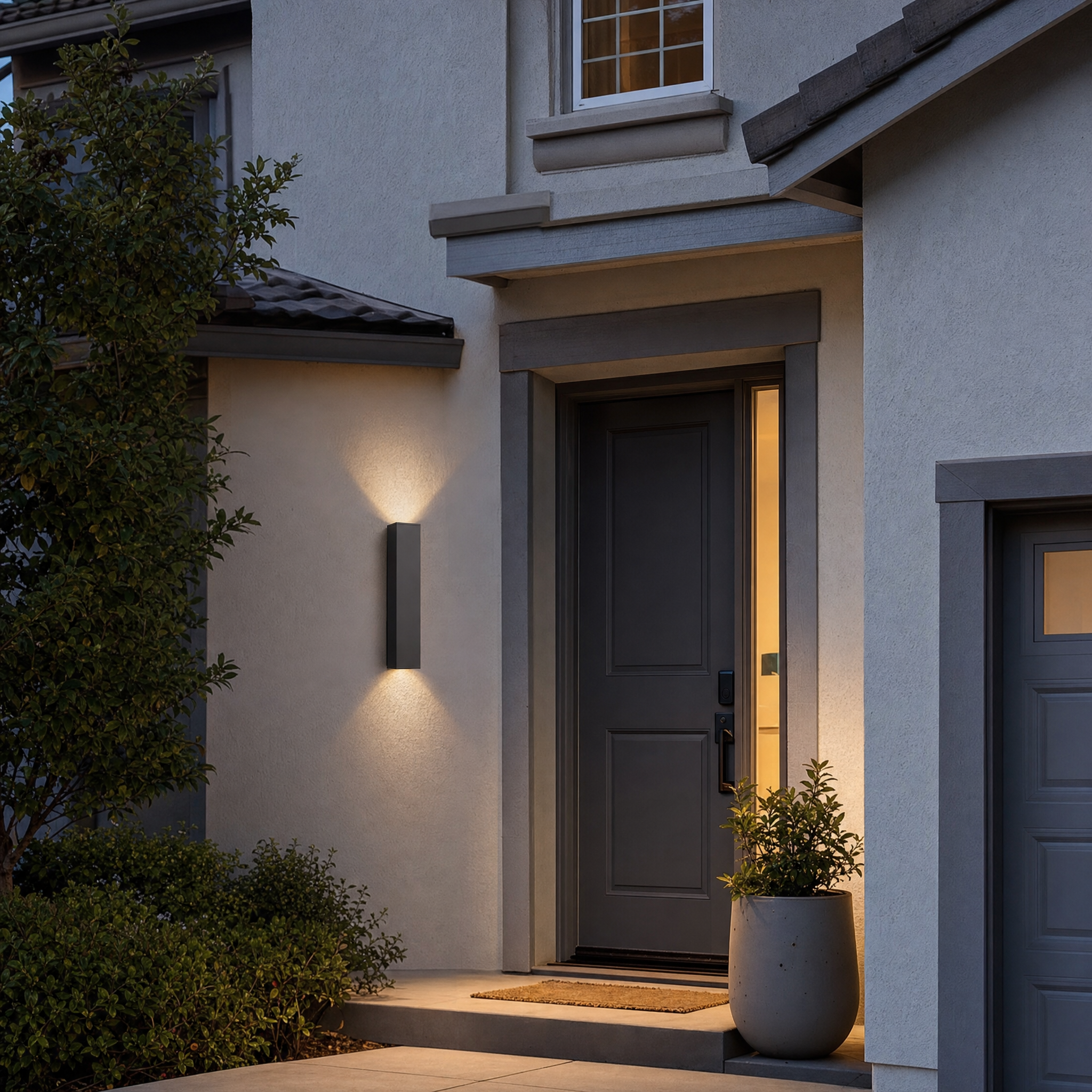 Charcoal up-and-down exterior wall light glowing beside a suburban front door at dusk